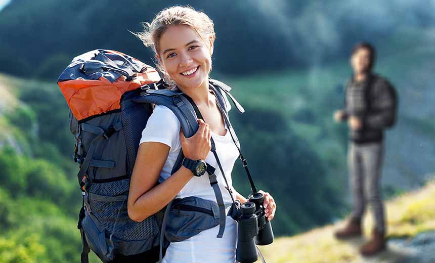 Woman with backpack over her shoulder smiling at the camera with with blurred hilly scene in the background 